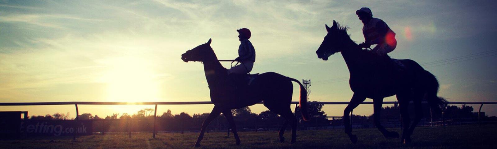 Two jockeys riding on the Fontwell Park Racecourse with the sunset behind them.
